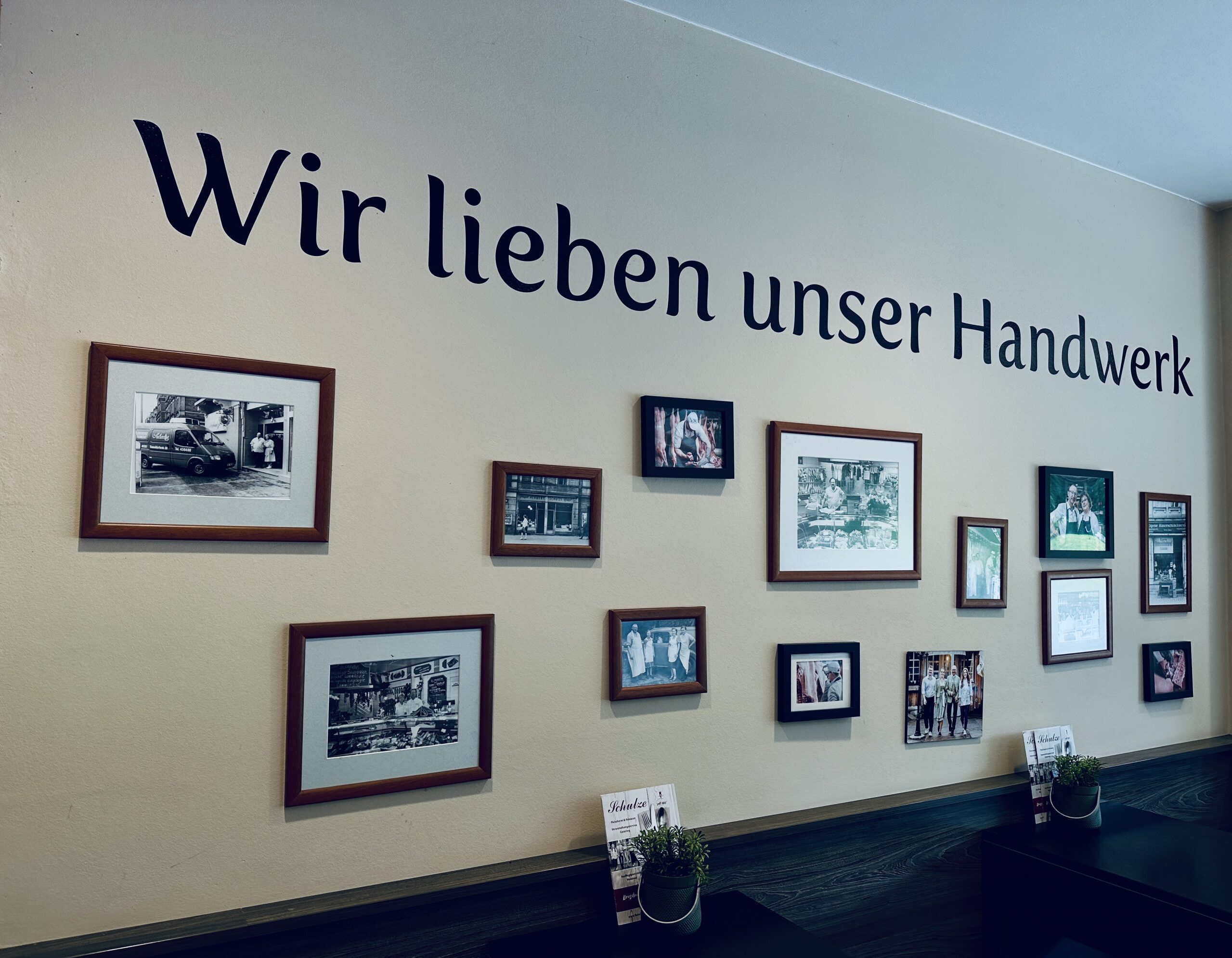 Fotowand in der Fleischerei Schulze in Dresden mit dem Schriftzug „Wir lieben unser Handwerk“ und gerahmten Bildern zur Unternehmensgeschichte.