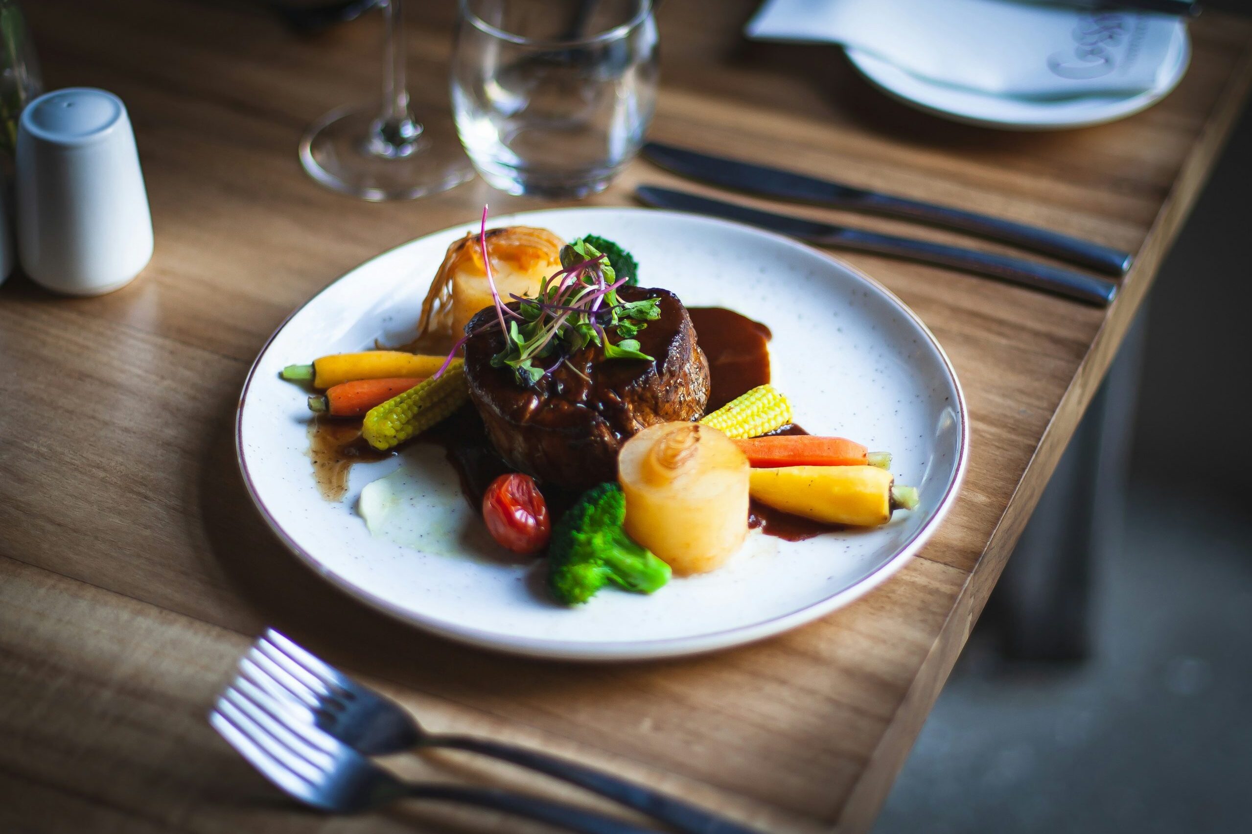 Angerichteter Hauptgang mit Fleisch in Sauce, Gemüse und Beilagen auf einem weißen Teller am Holztisch.